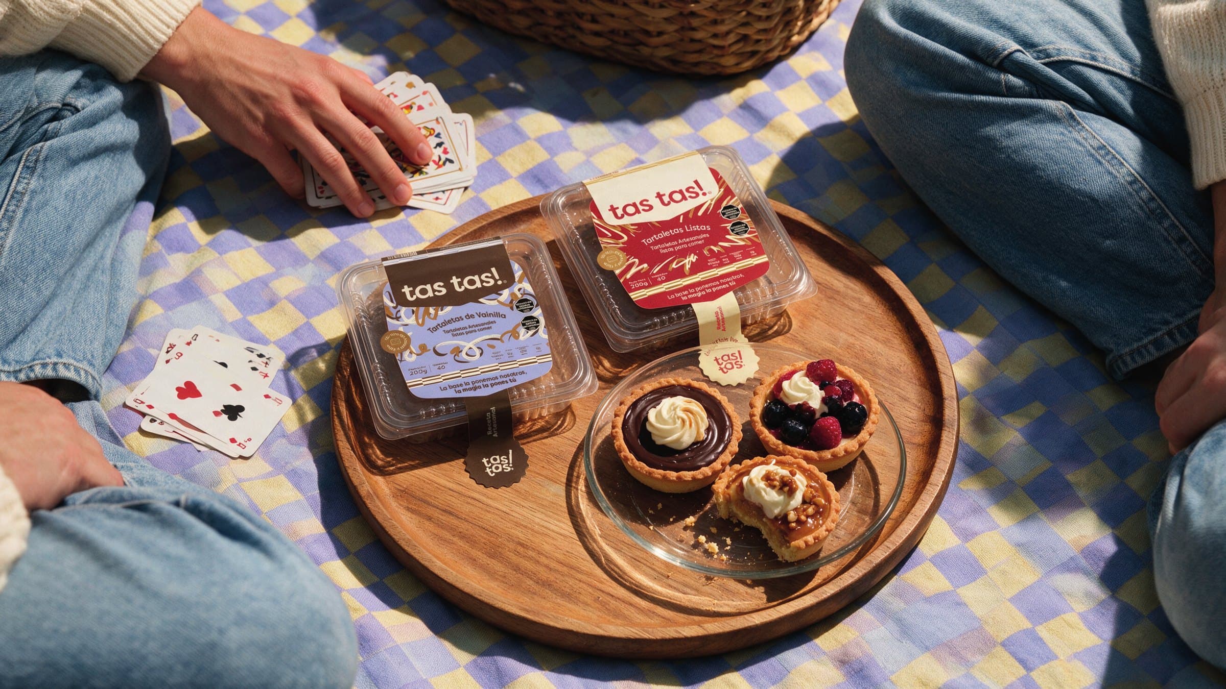 Wooden tray with two tas tas! packs and served tartlets at a picnic on a checked blanket